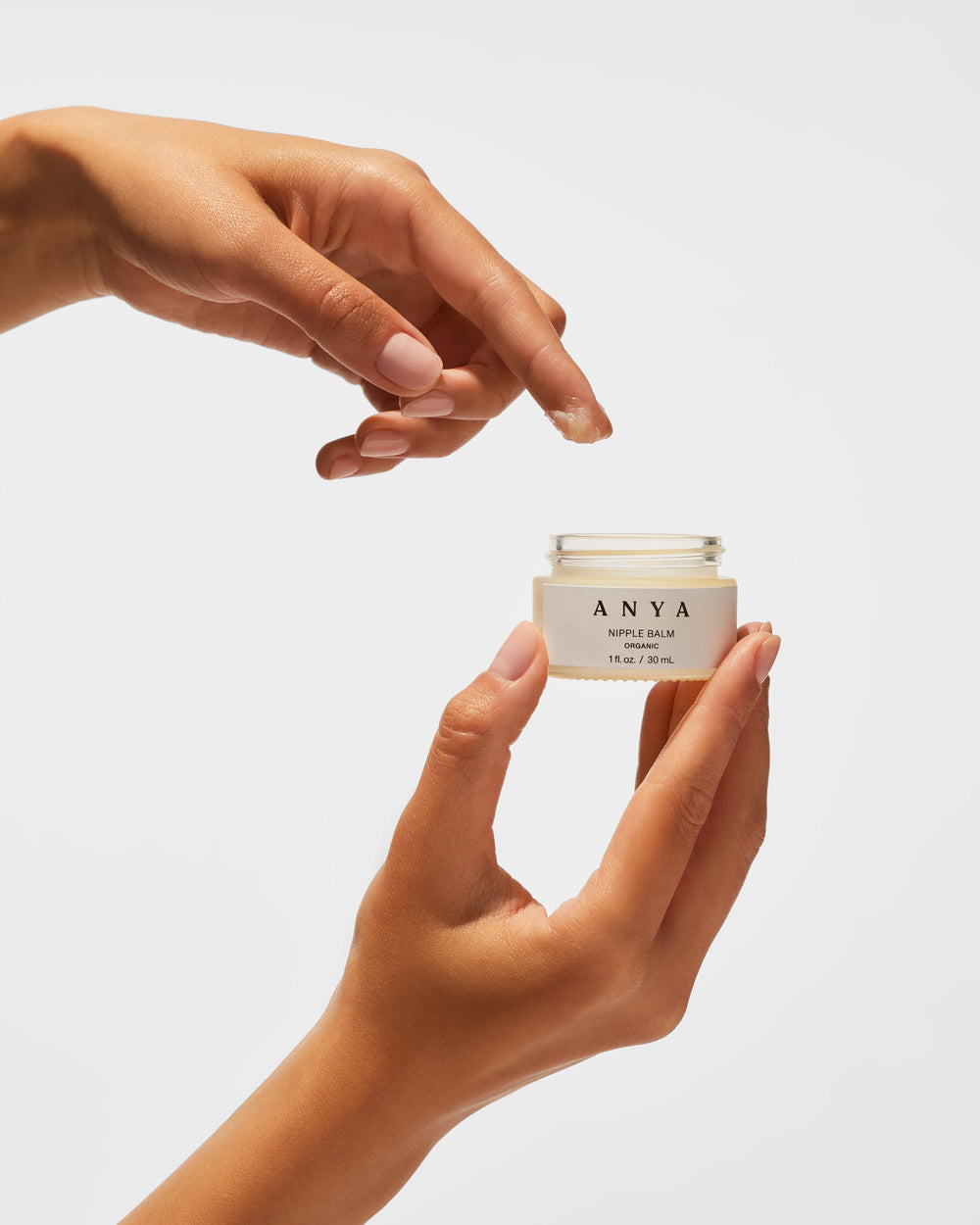 Two hands against a plain light gray background: one hand holds an open glass jar of "ANYA Nipple Balm, Organic, 1 fl. oz. / 30 mL" with a cream-colored label, while the other hand, positioned above, has a small dab of the balm on its extended index fingertip.